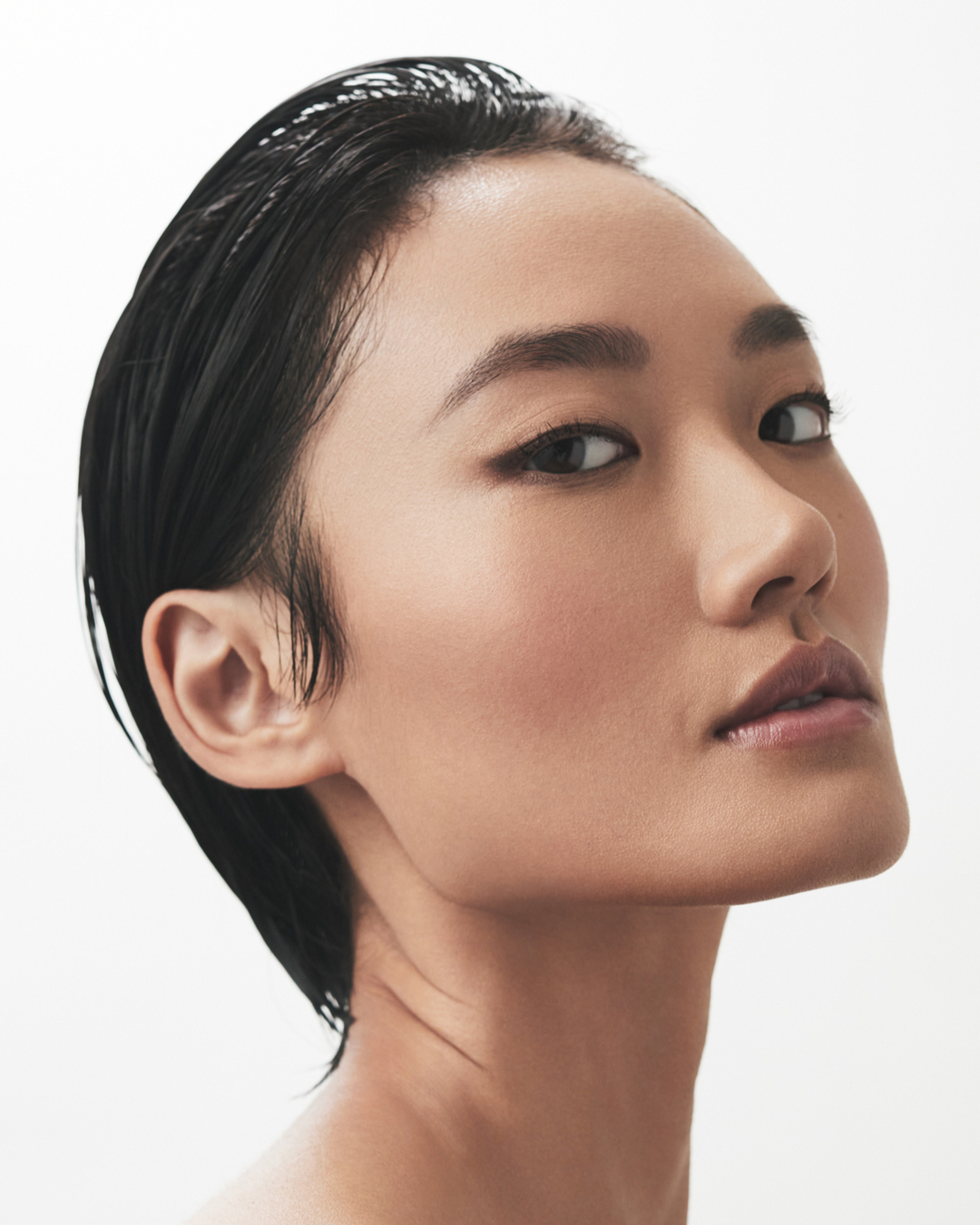 Close-up beauty shot of a young Asian woman with slicked-back black hair against a clean white backdrop, showcasing a natural makeup look.