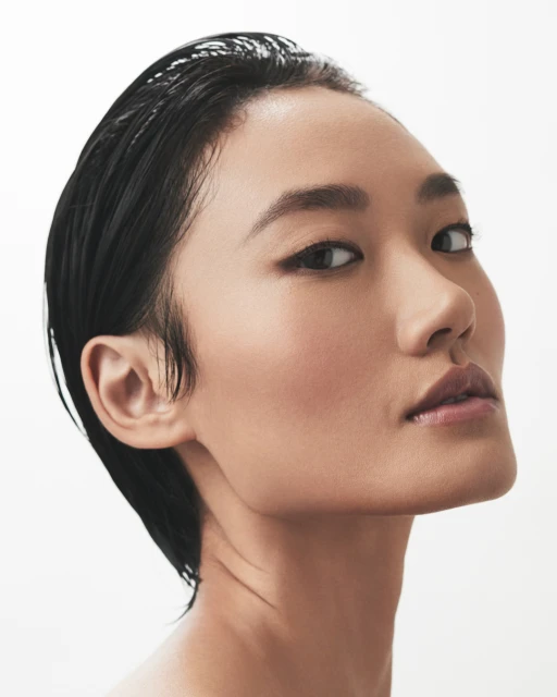 Close-up beauty shot of a young Asian woman with slicked-back black hair against a clean white backdrop, showcasing a natural makeup look.
