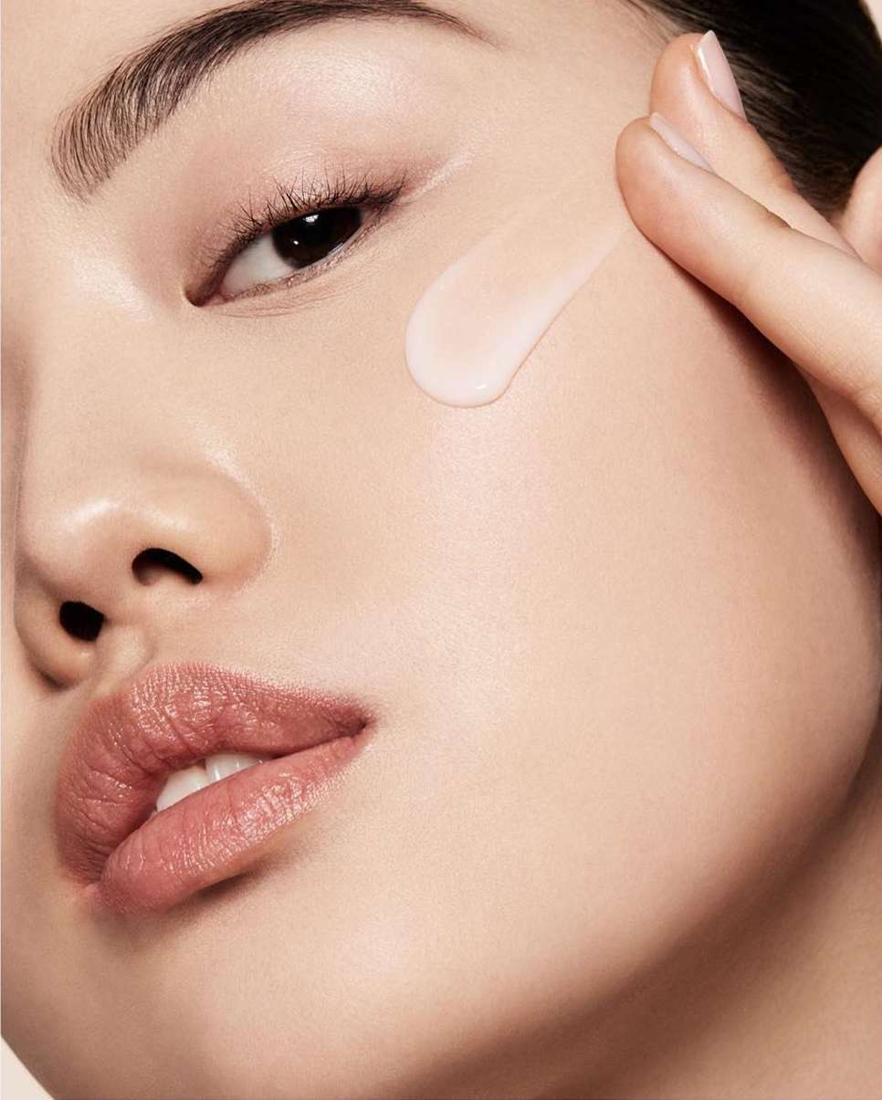 Close-up of an Asian woman's face with a drop of cream on her cheek being smoothed with a finger; skincare or beauty concept.