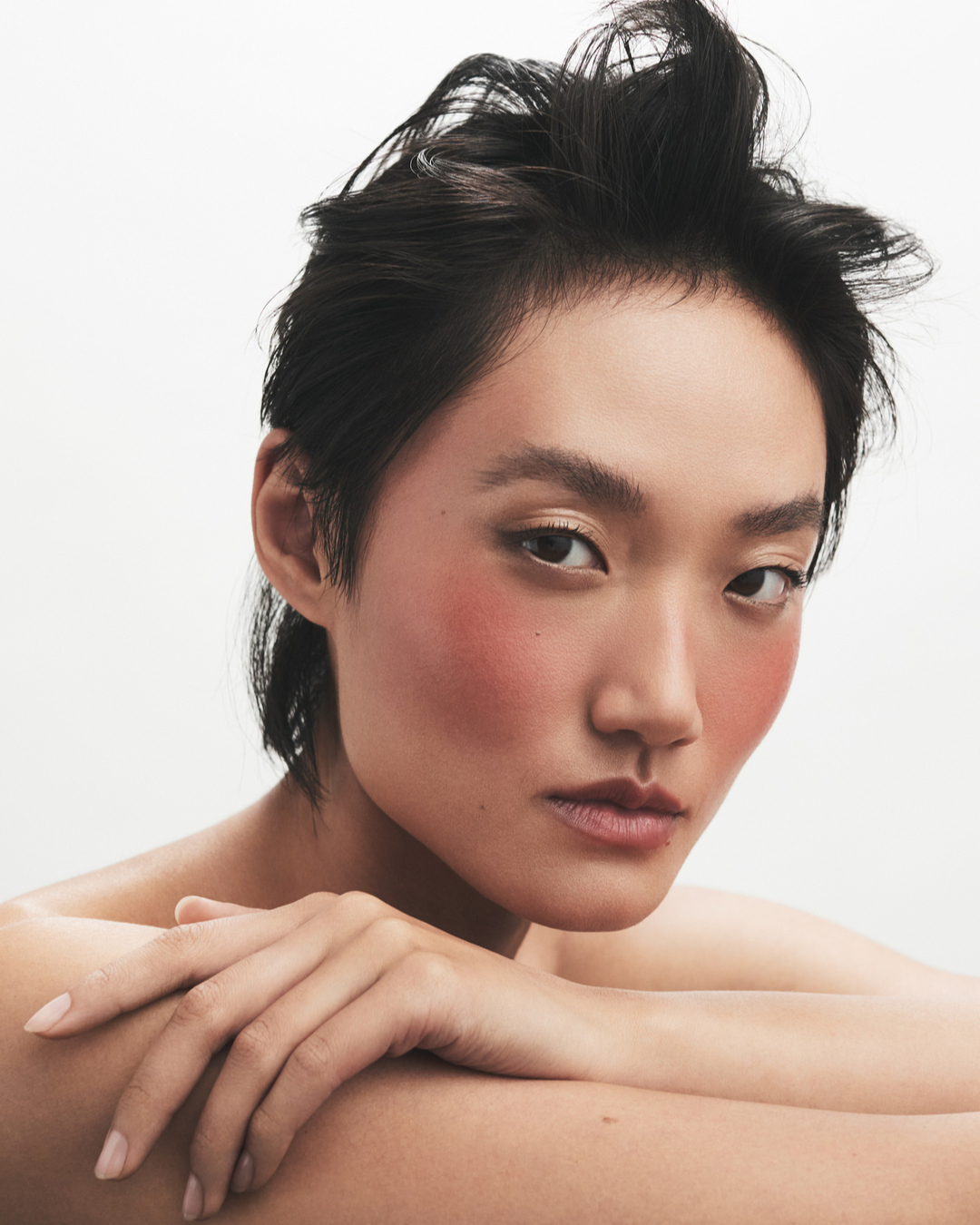 Close-up portrait of a young adult woman with dark hair and peach blush, posing in a photo studio with a bright white backdrop.