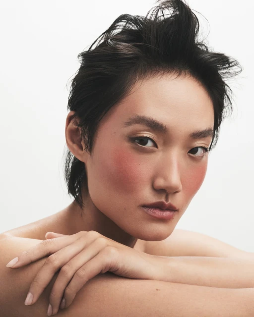 Close-up portrait of a young adult woman with dark hair and peach blush, posing in a photo studio with a bright white backdrop.