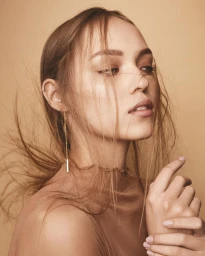 Portrait of a woman with long loose brown hair, wearing a neutral-toned top and thin drop earrings against a warm beige background.