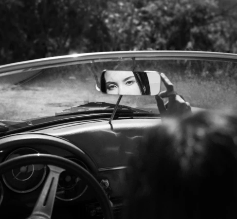 Black and white image shows a woman's eyes reflected in a car's rearview mirror, conveying a sense of nostalgia and introspection.
