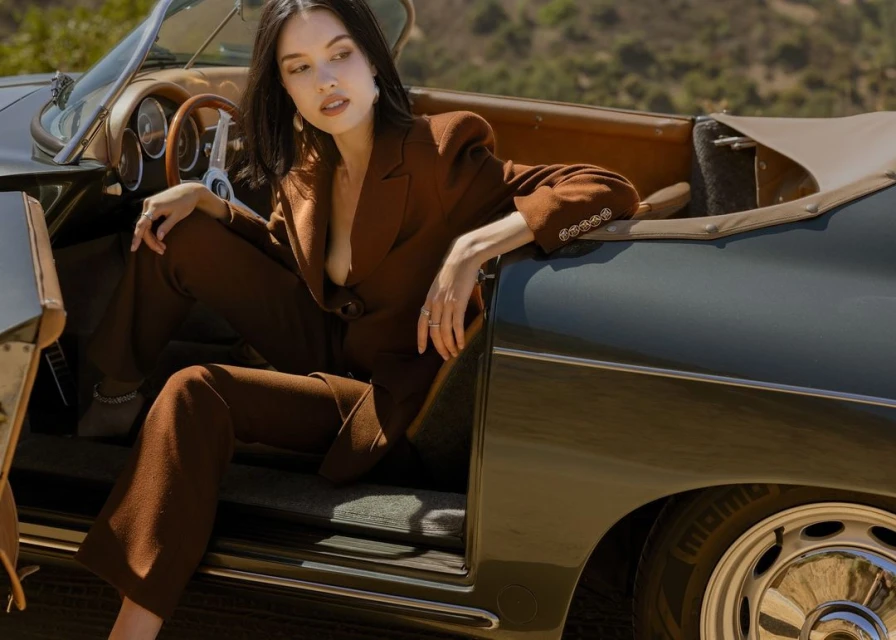 An adult female wearing a brown suit poses in a vintage convertible car with a neutral expression against an outdoor, natural background.