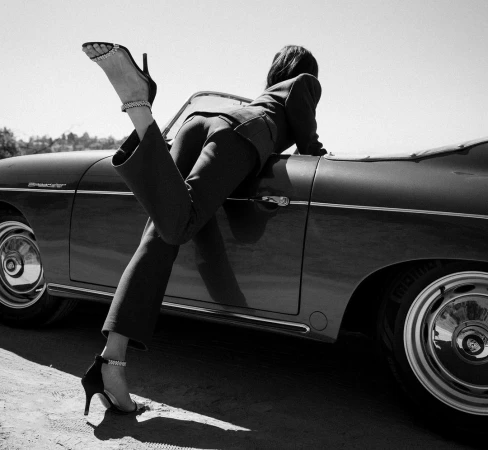 Woman in dark suit leaning over the side of a vintage convertible car with one leg kicked up high in a black and white photograph