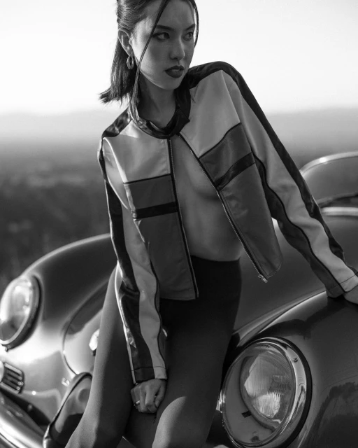 Stylish woman in racing jacket posing with classic car. Black and white image featuring fashion and automotive elements.