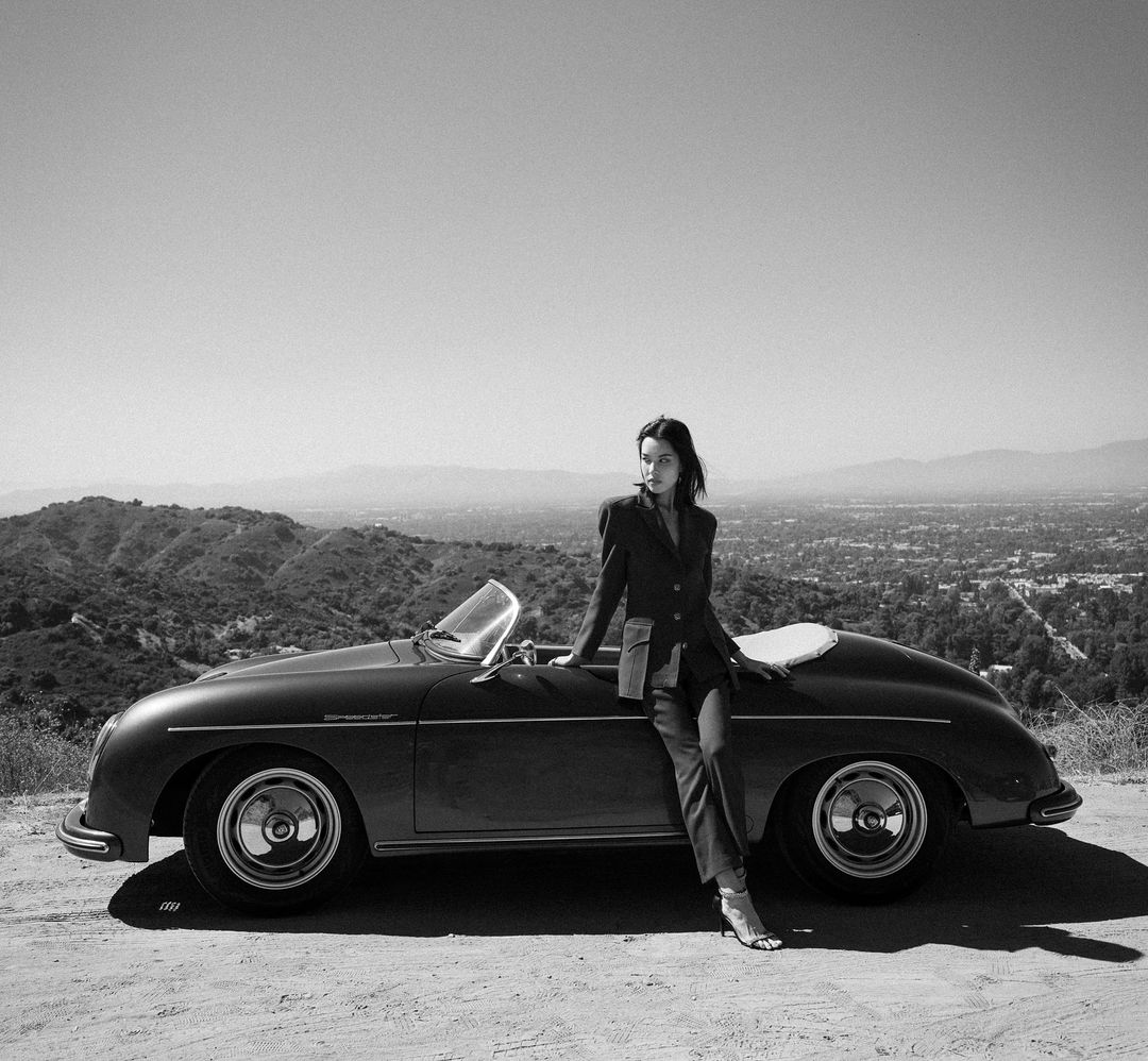 Woman in suit poses with a classic Porsche convertible car, set against a scenic landscape backdrop on a sunny day in black and white.