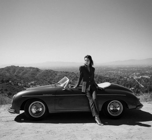 Woman in suit poses with a classic Porsche convertible car, set against a scenic landscape backdrop on a sunny day in black and white.