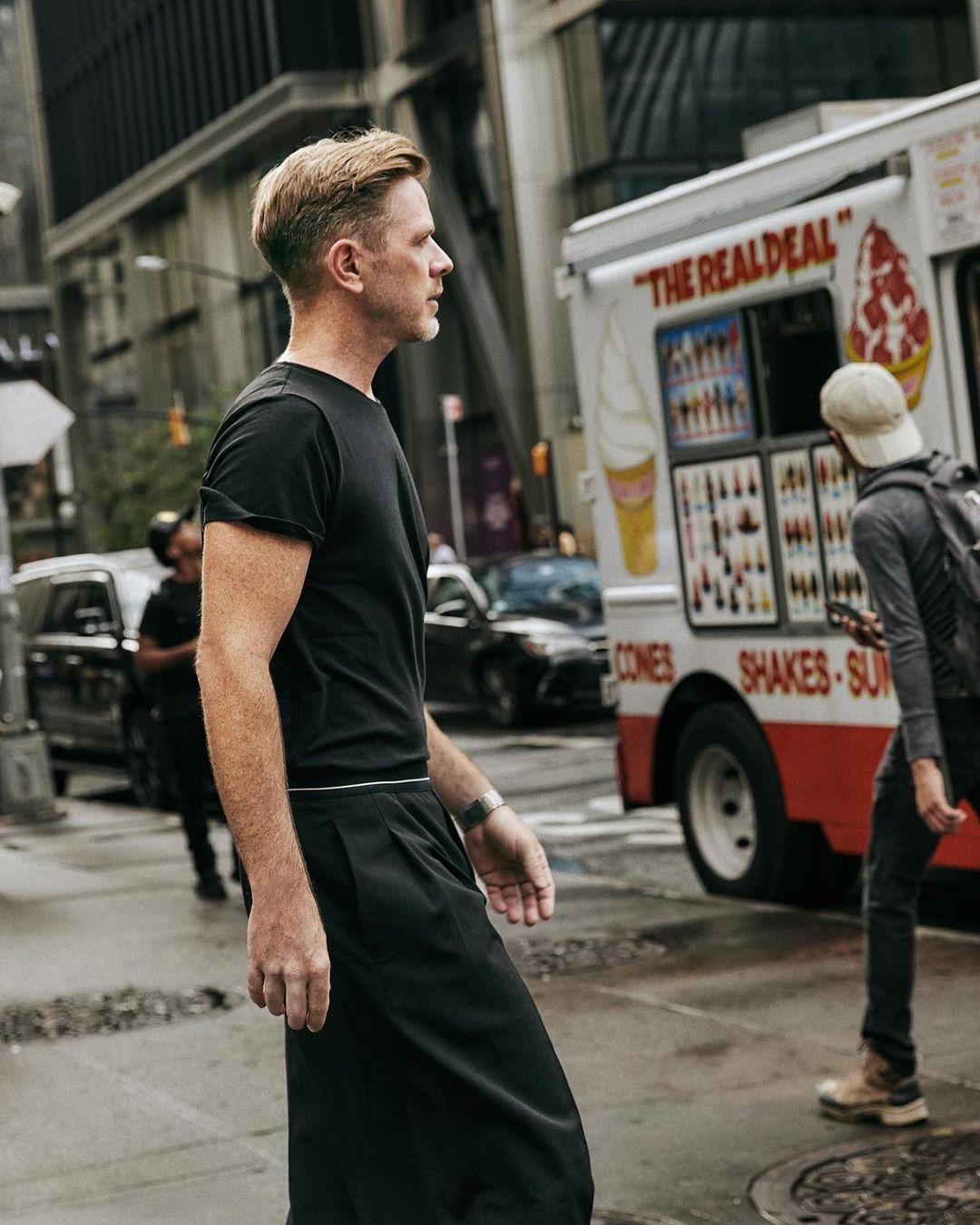 A fair-skinned man in black apparel walks in an urban setting, with an ice cream truck and modern buildings in the background.