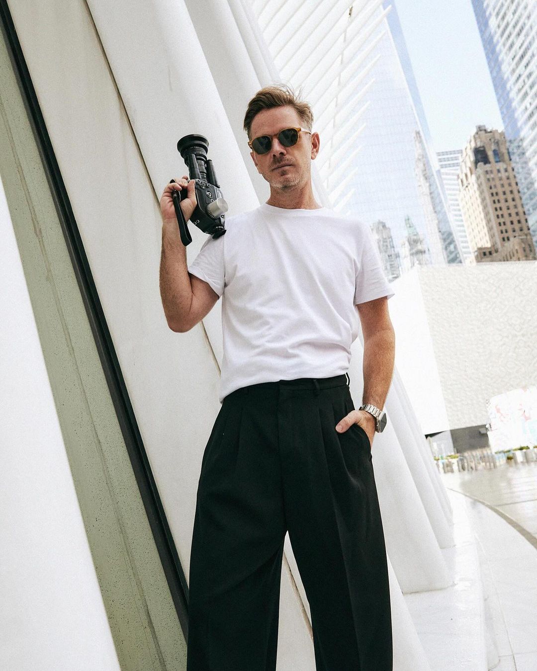 Adult male in sunglasses poses outdoors in city holding black camcorder with white shirt and dark slacks against modern architecture.