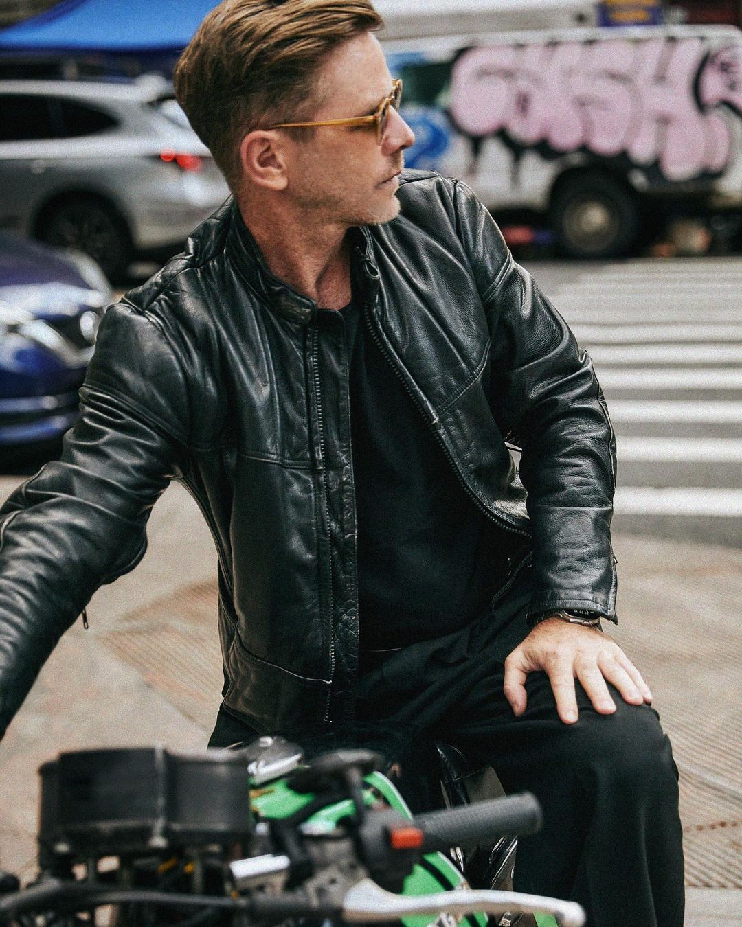 An adult male in a black leather jacket sits on a green Kawasaki motorcycle on a city street with cars and a graffiti-covered truck visible.