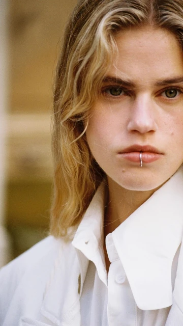 Close-up portrait of a fair-skinned person with blonde hair, wearing a white shirt and lip ring, gazing directly at the camera.