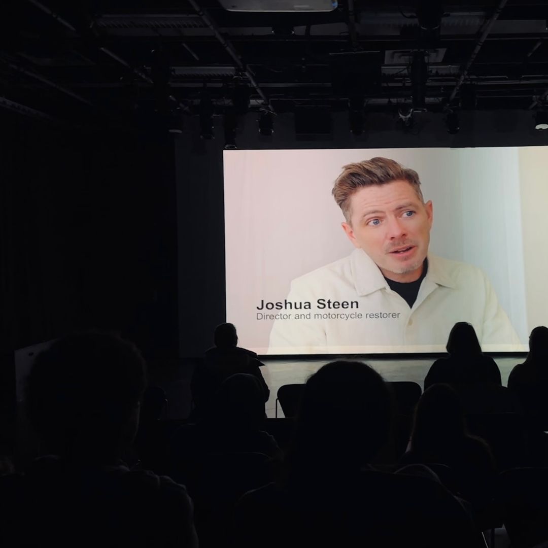 Audience watches speaker Joshua Steen on large screen in dark room. Overhead lights illuminate the presentation in an indoor setting.