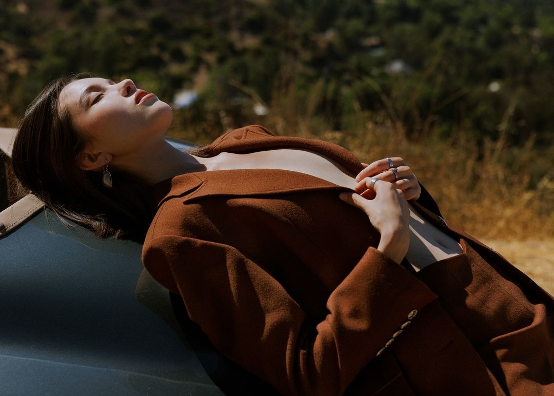 Woman in brown blazer reclining against a dark car with eyes closed, bathed in warm sunlight against a blurred natural background