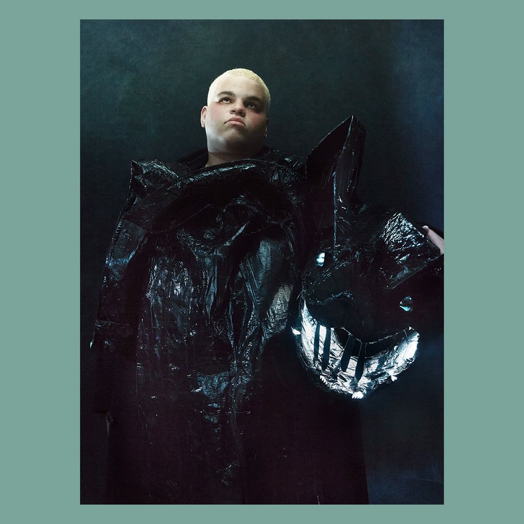 Portrait of an adult male with a shaved head wearing an elaborate black, shiny, plastic-like costume with a geometric smiling face.