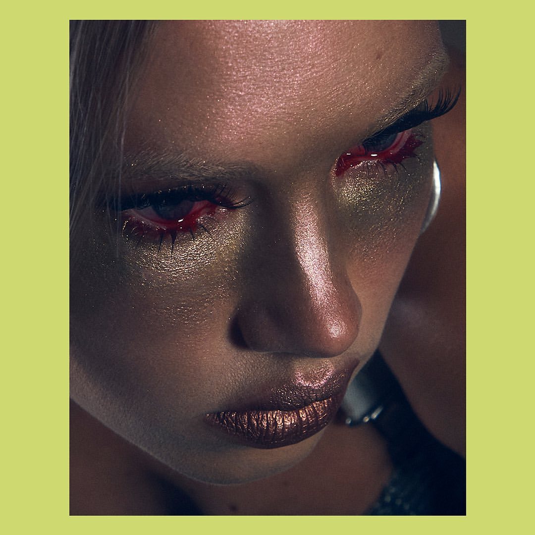 Dramatic close-up portrait of an adult female with distinctive metallic gold eye makeup and red detailing, set against a vibrant chartreuse background.