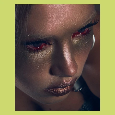 Dramatic close-up portrait of an adult female with distinctive metallic gold eye makeup and red detailing, set against a vibrant chartreuse background.