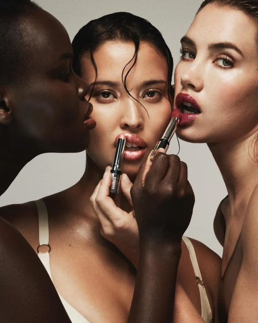 Three diverse women pose closely, applying lipstick. Beauty shot with high-key lighting showcases makeup and skincare for a cosmetics campaign.
