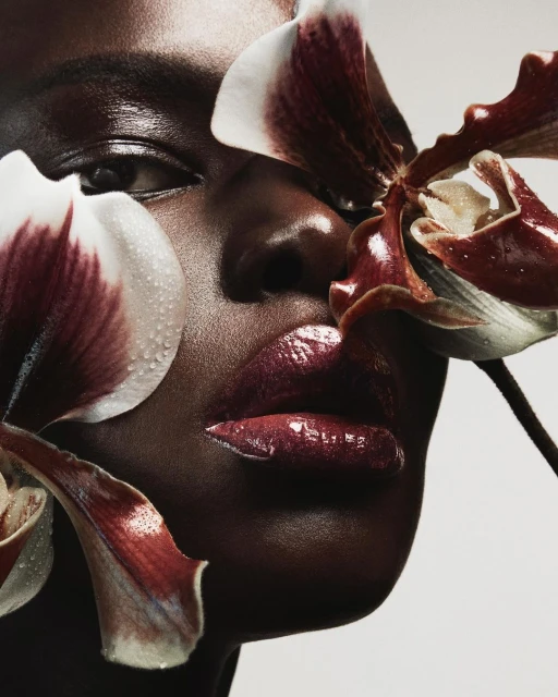 Close-up of a Black woman with glossy lips, surrounded by orchid flowers, showcasing beauty and skincare in a studio setting.