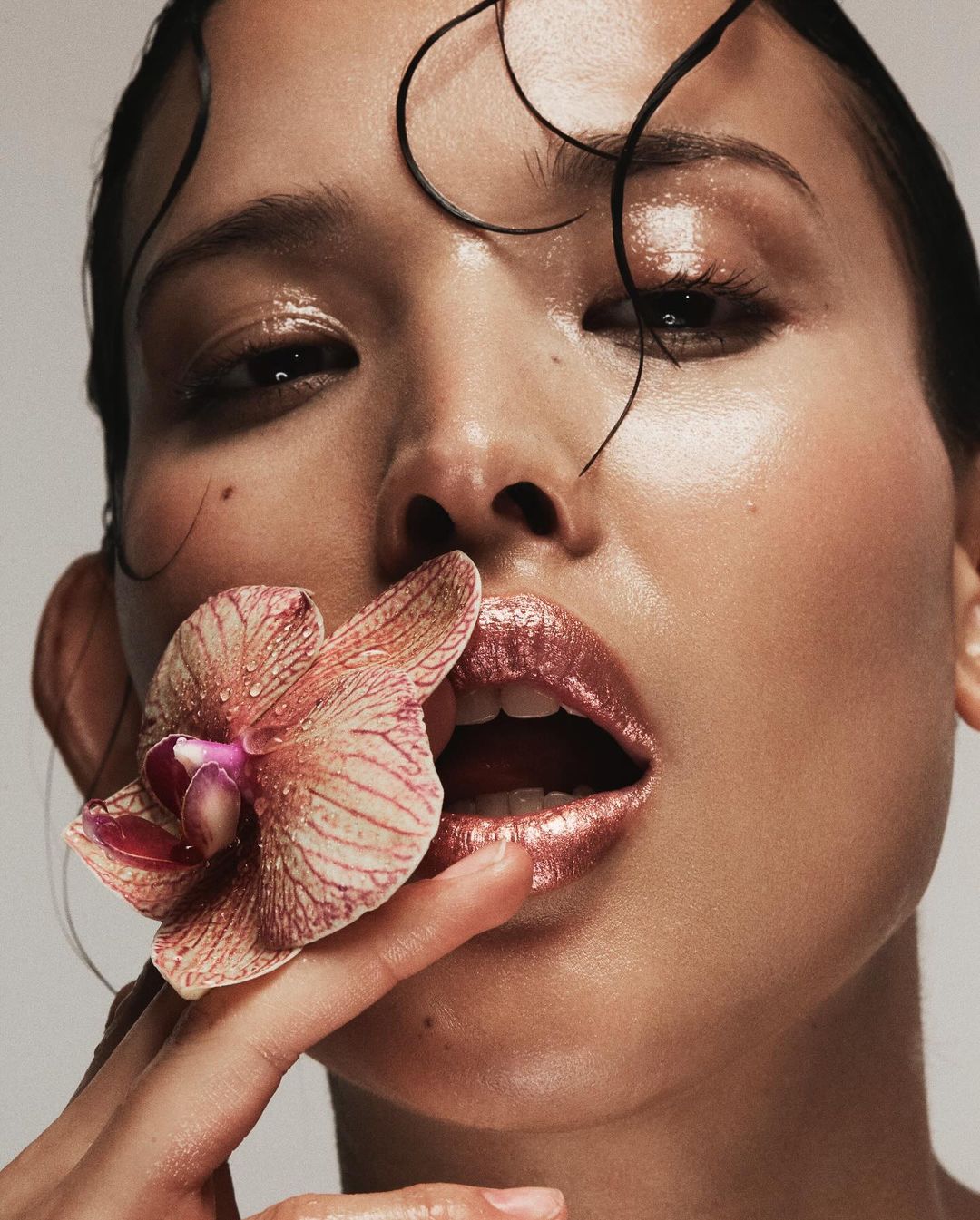 Close-up of a person with wet hair holding an orchid near their mouth, featuring glossy lips and soft, diffused lighting.