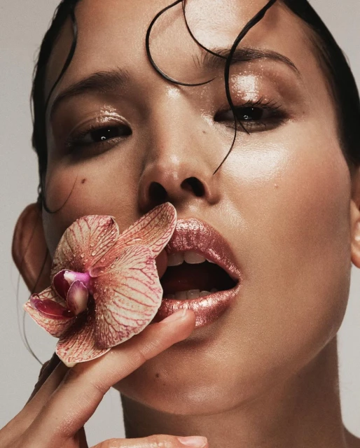 Close-up of a person with wet hair holding an orchid near their mouth, featuring glossy lips and soft, diffused lighting.