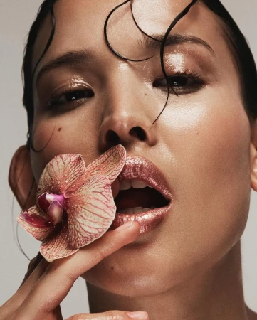 Close-up of a person with wet hair holding an orchid near their mouth, featuring glossy lips and soft, diffused lighting.