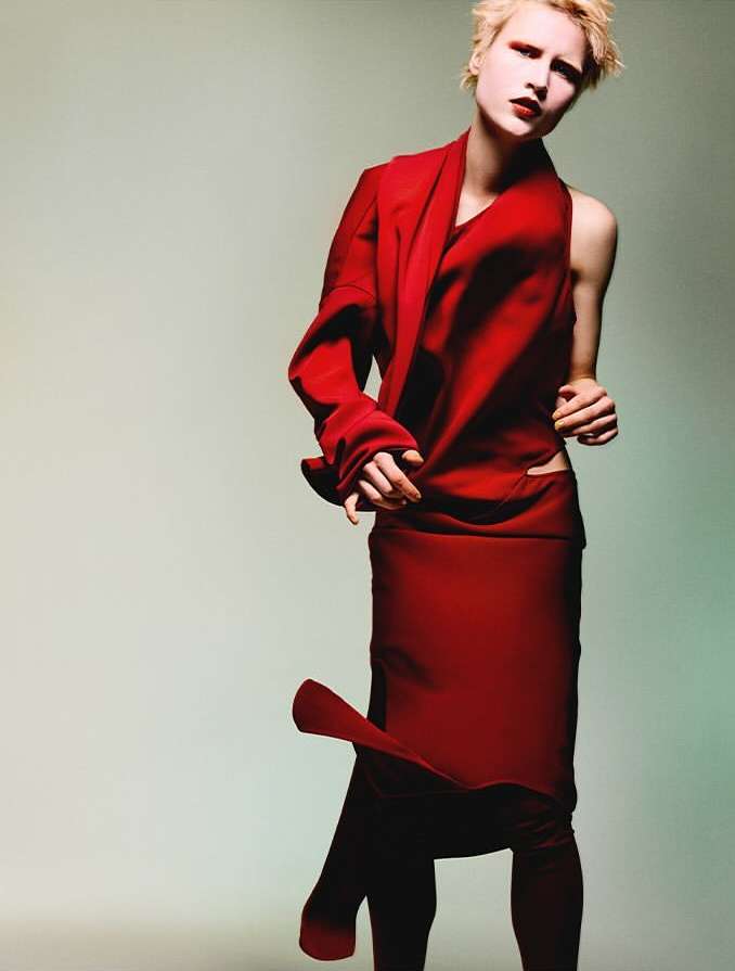 A model in a vibrant red outfit poses indoors with dramatic makeup in a fashion editorial shot, set against a neutral backdrop.