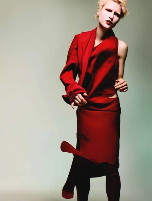 A model in a vibrant red outfit poses indoors with dramatic makeup in a fashion editorial shot, set against a neutral backdrop.