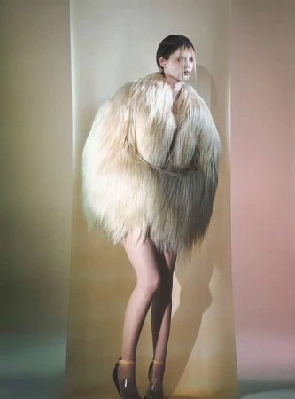 An adult female model poses in a creamy, full-length shaggy fur coat and clear high heels in a fashion studio with a gradient backdrop.