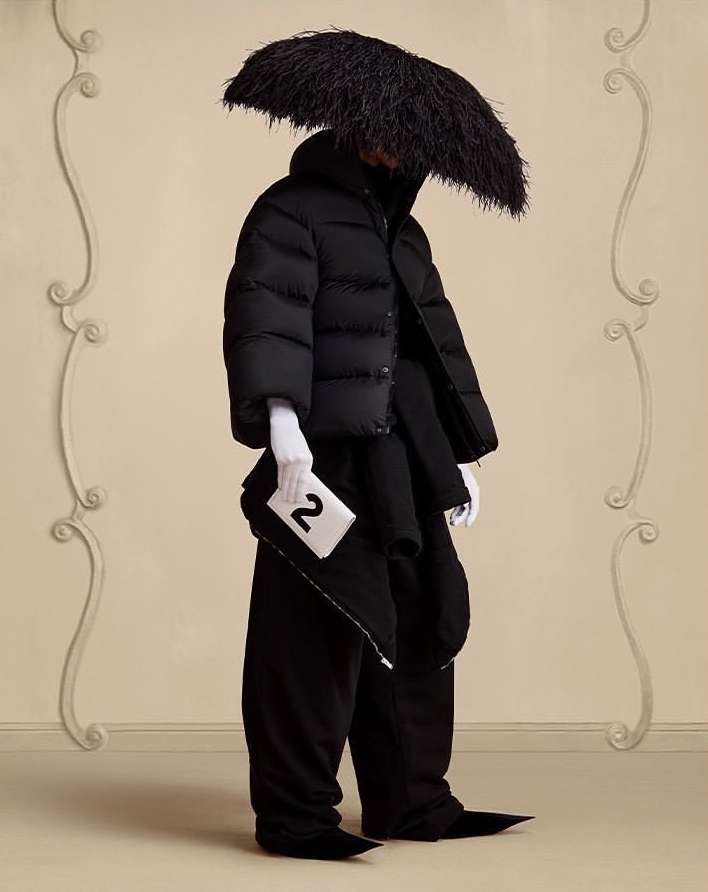 Fashionable individual in avant-garde black puffer jacket, wide trousers, white gloves, and feathered headwear against a neutral backdrop.