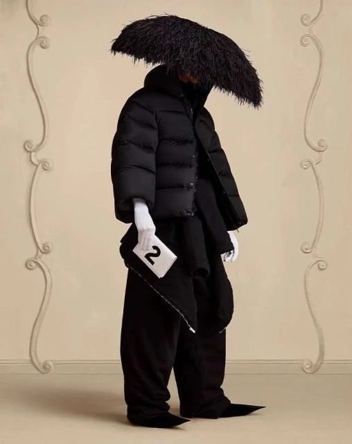 Fashionable individual in avant-garde black puffer jacket, wide trousers, white gloves, and feathered headwear against a neutral backdrop.