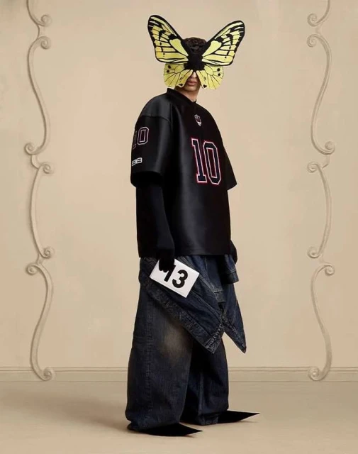 Person in oversized streetwear fashion wearing a butterfly mask, baggy jeans, and pointy shoes against a neutral wall with moulding.