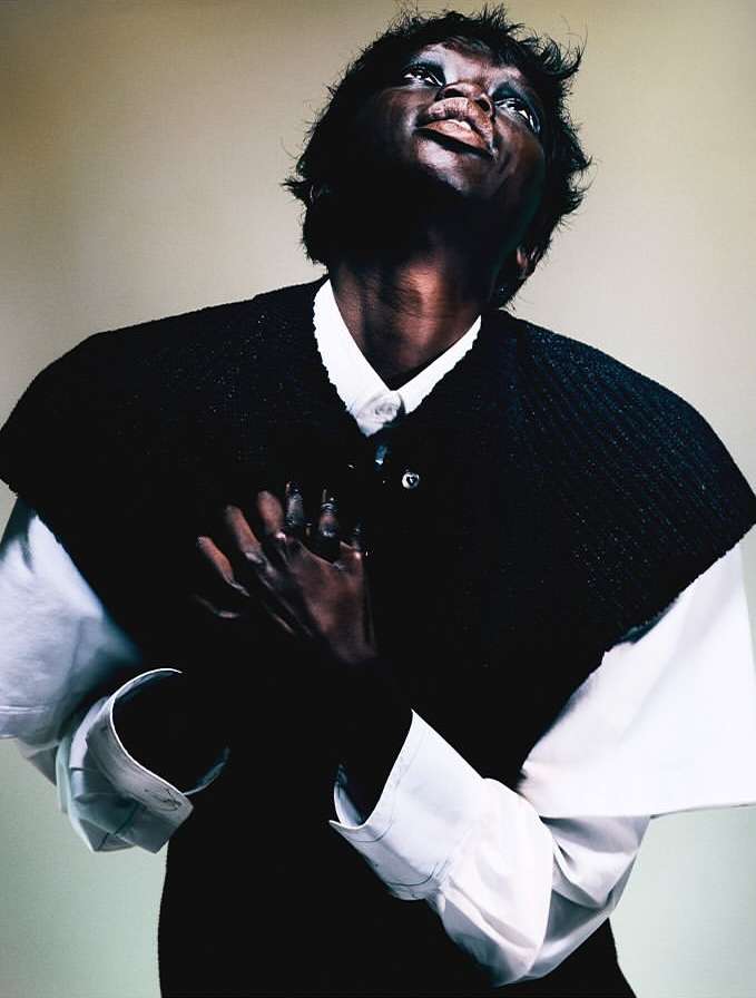 Portrait of an adult woman in a white collared shirt and dark vest looking up with clasped hands in a photo studio.