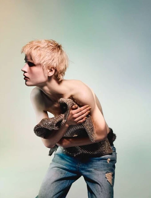 Bare-chested model with light blond hair poses in ripped jeans, holding a metallic fabric against a gradient background in a studio shot.