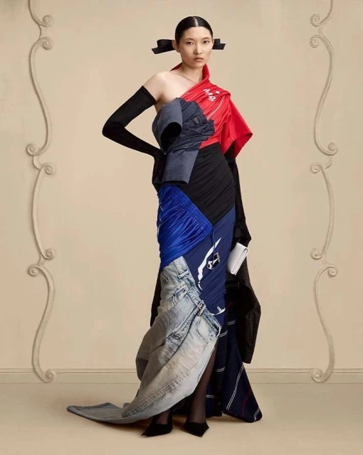Fashion model poses in a full-length shot, wearing a unique dress made of repurposed clothing materials, in a studio setting.