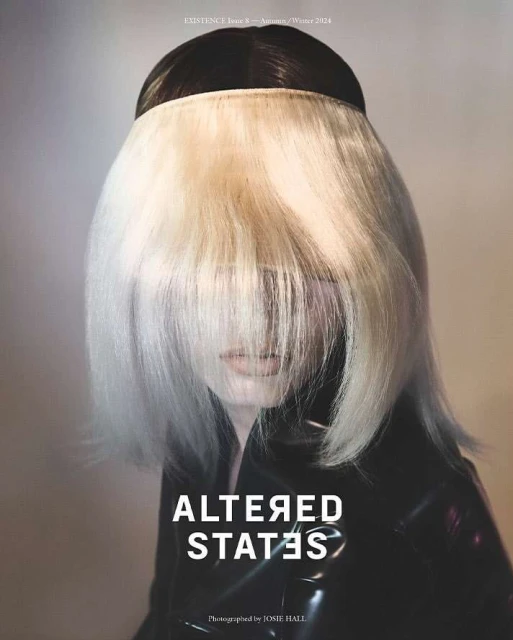 Fashion shoot featuring a person with black and blonde hair wearing a black shiny jacket, shot in a studio with a neutral backdrop.