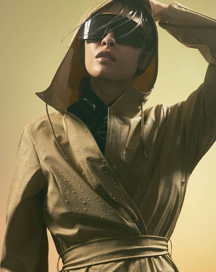 Woman in futuristic sunglasses and tan raincoat with hood up, standing in a studio against a yellow gradient background.