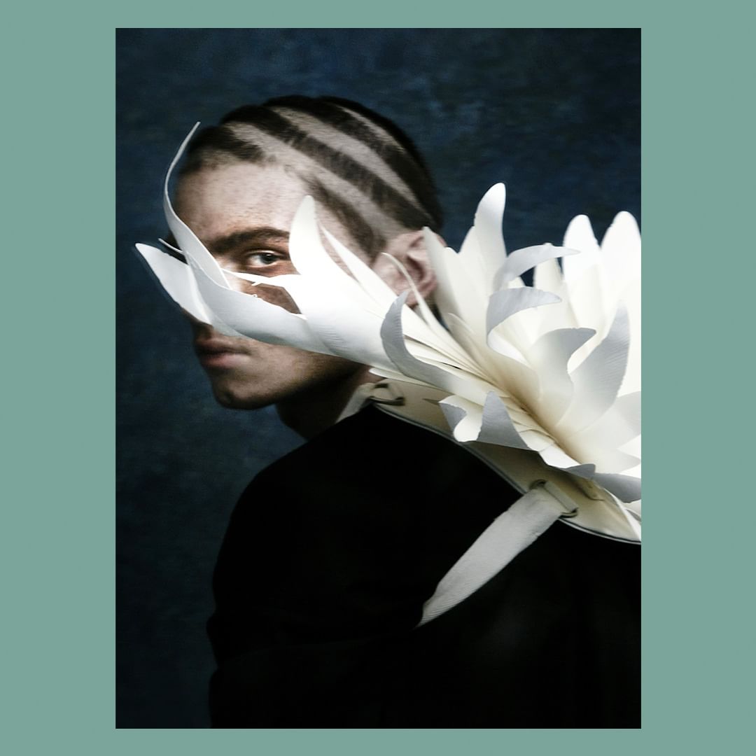 Portrait of a fair-skinned person with shaved striped hair and a black top; a large white paper flower adorns their shoulder and face.