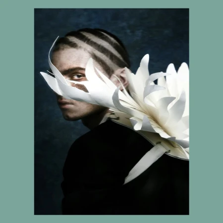 Portrait of a fair-skinned person with shaved striped hair and a black top; a large white paper flower adorns their shoulder and face.