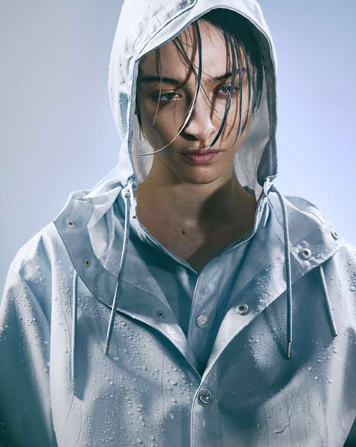 Close-up studio shot of a woman with dark hair wearing a blue hooded, button-down rain jacket, looking directly at the camera.