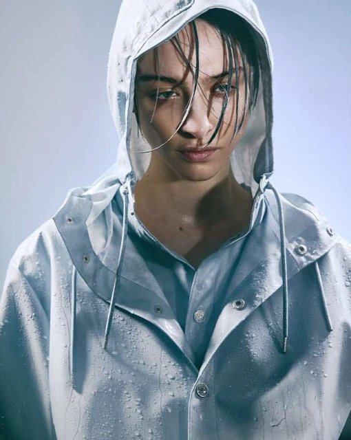 Close-up studio shot of a woman with dark hair wearing a blue hooded, button-down rain jacket, looking directly at the camera.
