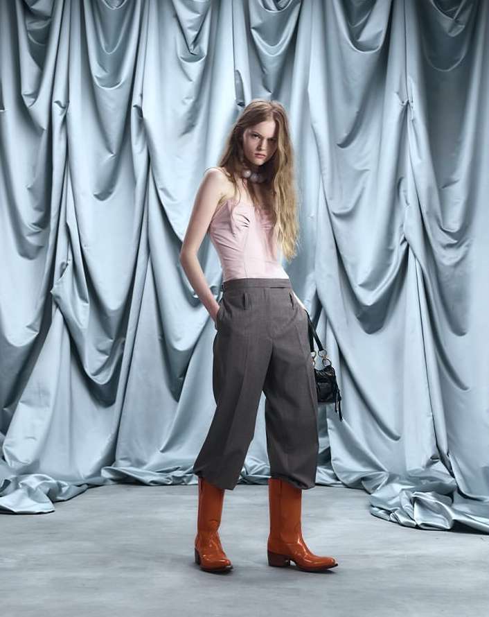 Full shot of a model in a pink top, grey culottes, and orange boots posing against a draped light blue fabric backdrop in a studio.
