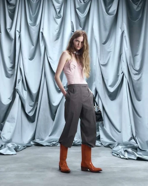 Full shot of a model in a pink top, grey culottes, and orange boots posing against a draped light blue fabric backdrop in a studio.