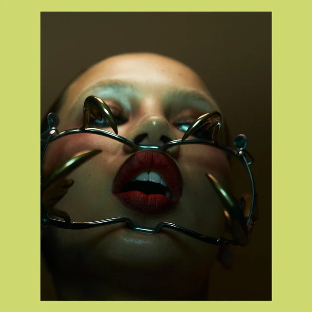 Dramatic beauty shot of a woman with bold red lipstick, an open mouth, and distinctive metallic face jewelry against a yellow backdrop.
