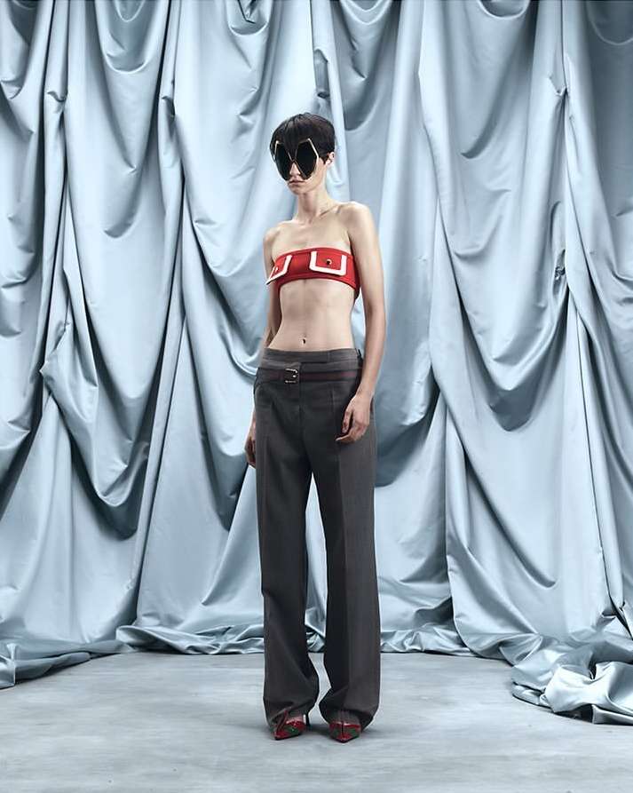 A woman in a red tube top and grey trousers stands in front of a draped blue fabric backdrop, captured in a studio fashion shot.