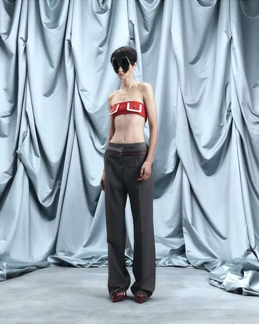 A woman in a red tube top and grey trousers stands in front of a draped blue fabric backdrop, captured in a studio fashion shot.