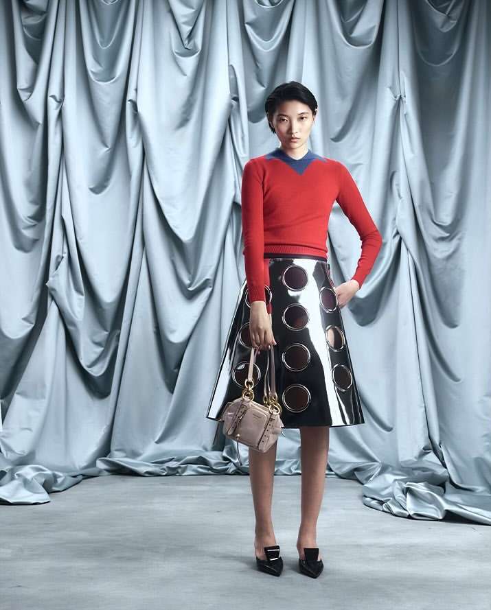 A striking studio shot showcases an Asian woman in a red sweater and black skirt, holding a handbag, set against a draped silver backdrop.