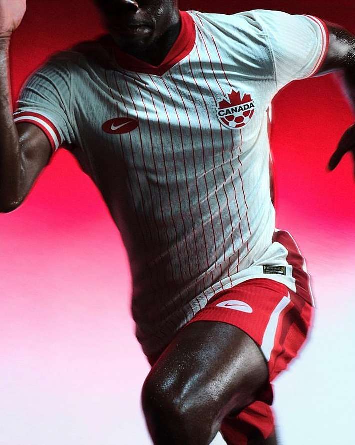 Athletic adult male in a grey Canada soccer jersey running against a vibrant red background, showcasing fitness and sports fashion.