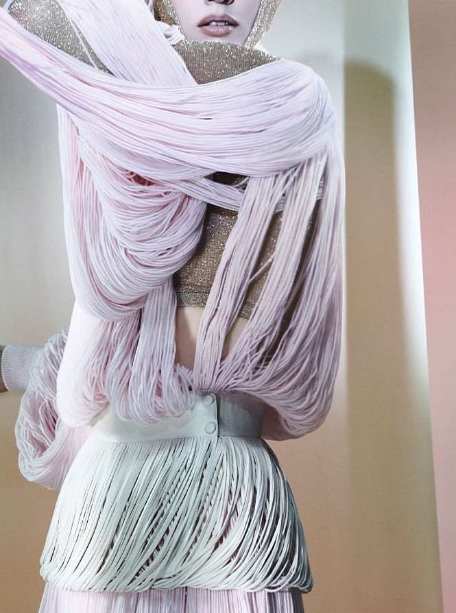 Fashion shot of a person in a beige interior wearing a gold top and a skirt and wrap made of long pink and white fringe.