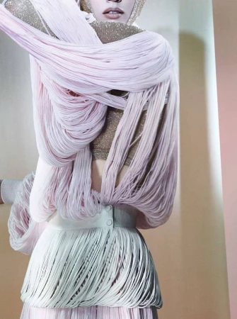 Fashion shot of a person in a beige interior wearing a gold top and a skirt and wrap made of long pink and white fringe.